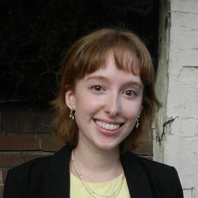 A person with short light auburn hair and straight bangs, wearing a black blazer over a light yellow top, layered necklaces, and dangling earrings.